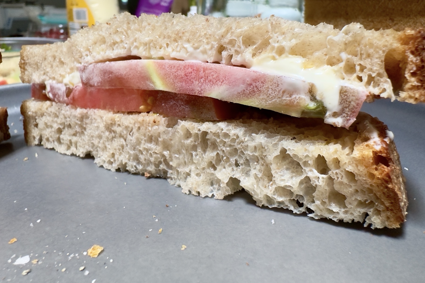 An up close view of a tomato sandwich. Two slices of homemade whole wheat sourdough sandwich bread around a slice of tomato dressed with mayonnaise, salt, and a little black pepper. It is cut on the diagonal, just as sandwiches should be. The plate is gray.