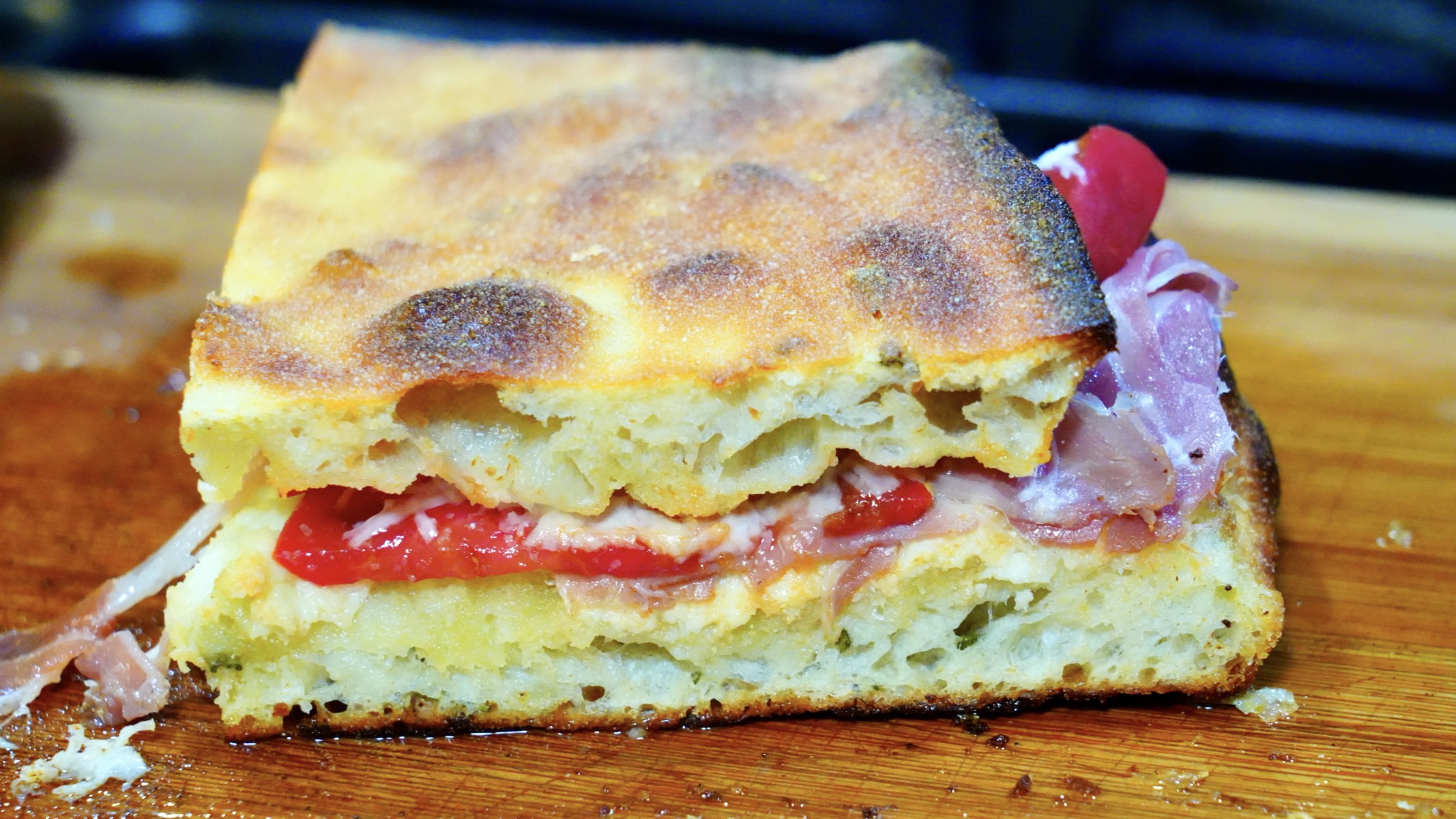 A fresh sandwich sits on a cutting board. The top is freshly baked crispy focaccia. The filling is prosciutto, fresh sliced tomatoes from the garden that are so so good, and melted pecorino cheese. The bottom slice is full of bubbles and looks amazing.