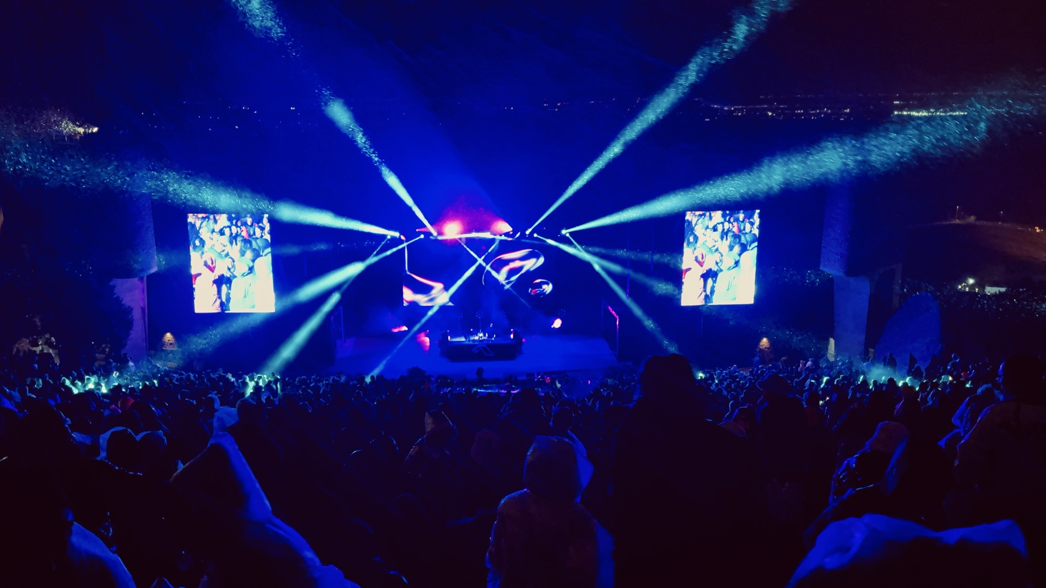 A concert crowd light with electric blue, the DJs are small on the distance stage, and snow is visible in the sweeping light show spots.